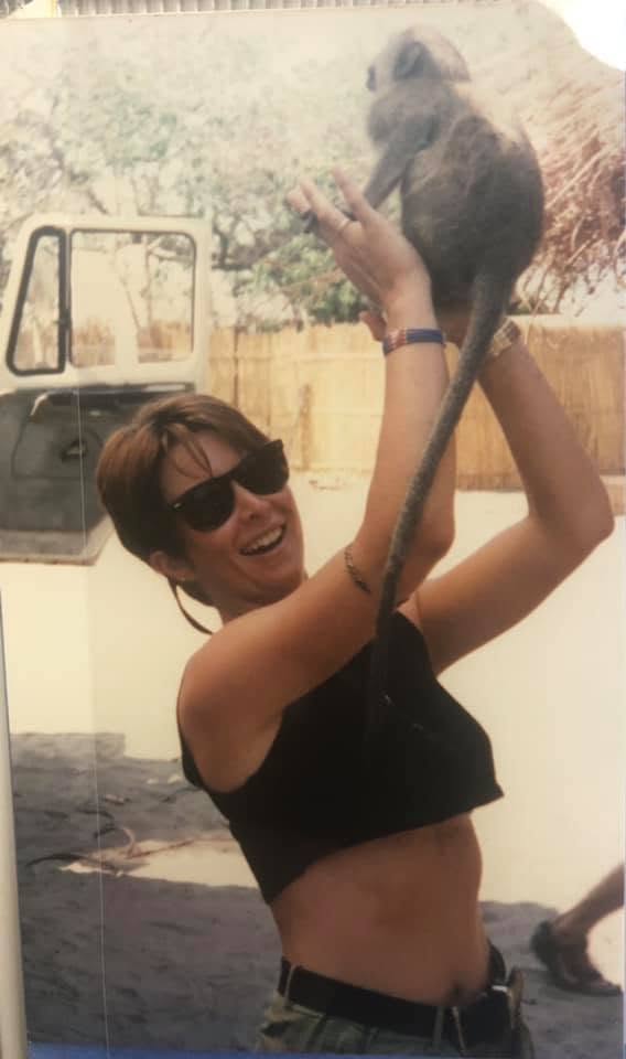 A woman wearing sunglasses smiles while lifting a monkey outdoors, with a vehicle and a natural background.