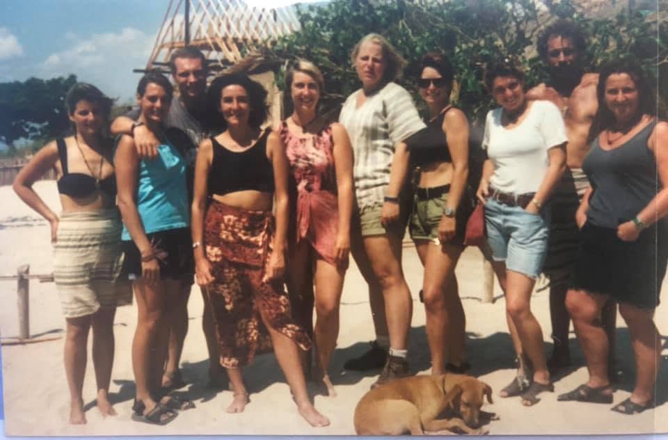 A group of ten individuals posing together on a sandy beach, with a dog lying on the ground. The people are dressed in casual summer attire, and the background features greenery and a wooden structure.