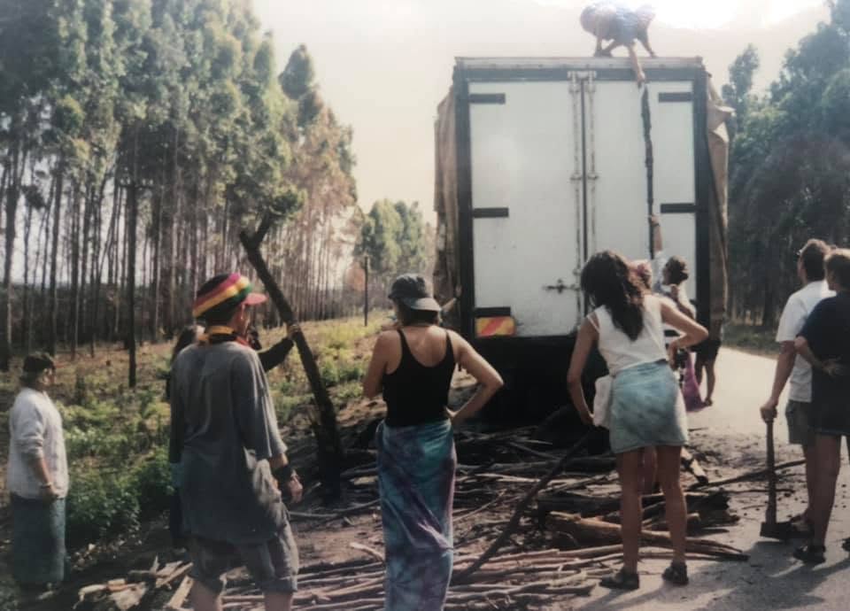 A group of people in casual clothing gathered around a truck on a dirt road, surrounded by trees. One person is climbing the truck, while others are observing and holding wooden sticks.