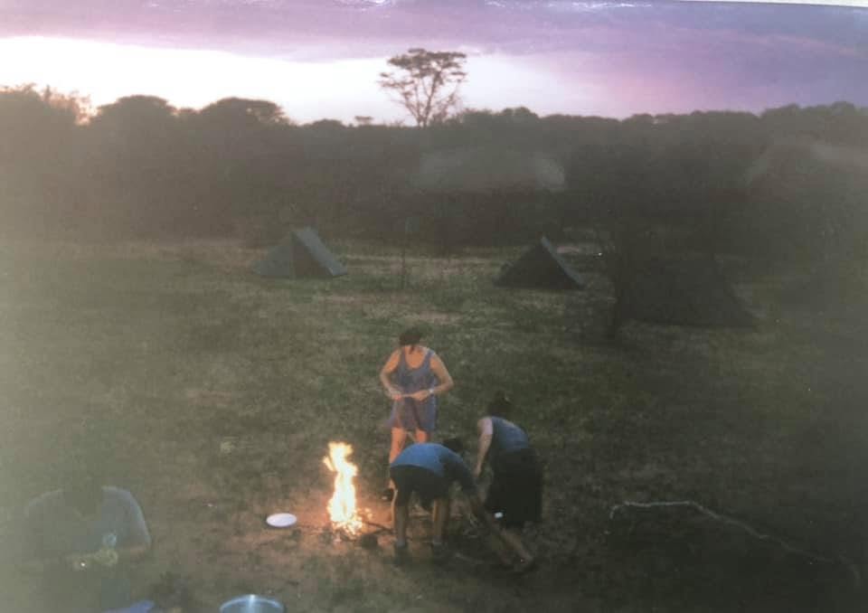 A campsite at dusk with four people gathered around a fire, one person stands while others are kneeling near the flames. In the background, several tents are set up against a twilight sky.