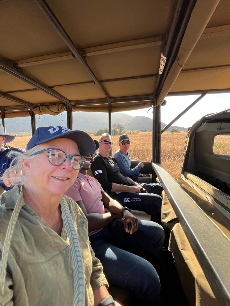 A group of four people sitting inside a safari vehicle, enjoying an outdoor adventure. The landscape features golden grass and hills in the background.