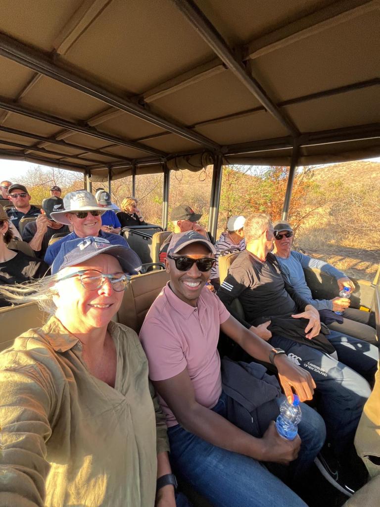 A group of people enjoying a safari ride in an open vehicle, with two individuals smiling in the foreground, surrounded by others in casual attire.