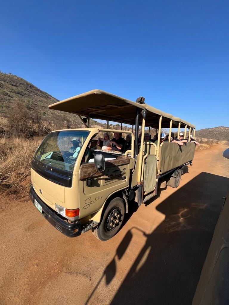 An open-top safari vehicle parked on a dirt road surrounded by sparse vegetation and hills under a clear blue sky.