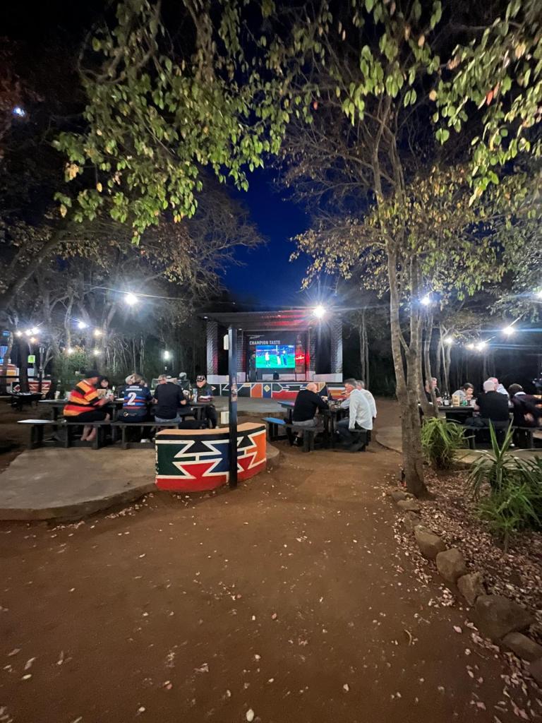 Outdoor viewing area at night with a large screen displaying a sports event, surrounded by trees and illuminated by string lights. Patrons seated at colorful tables enjoying the atmosphere.