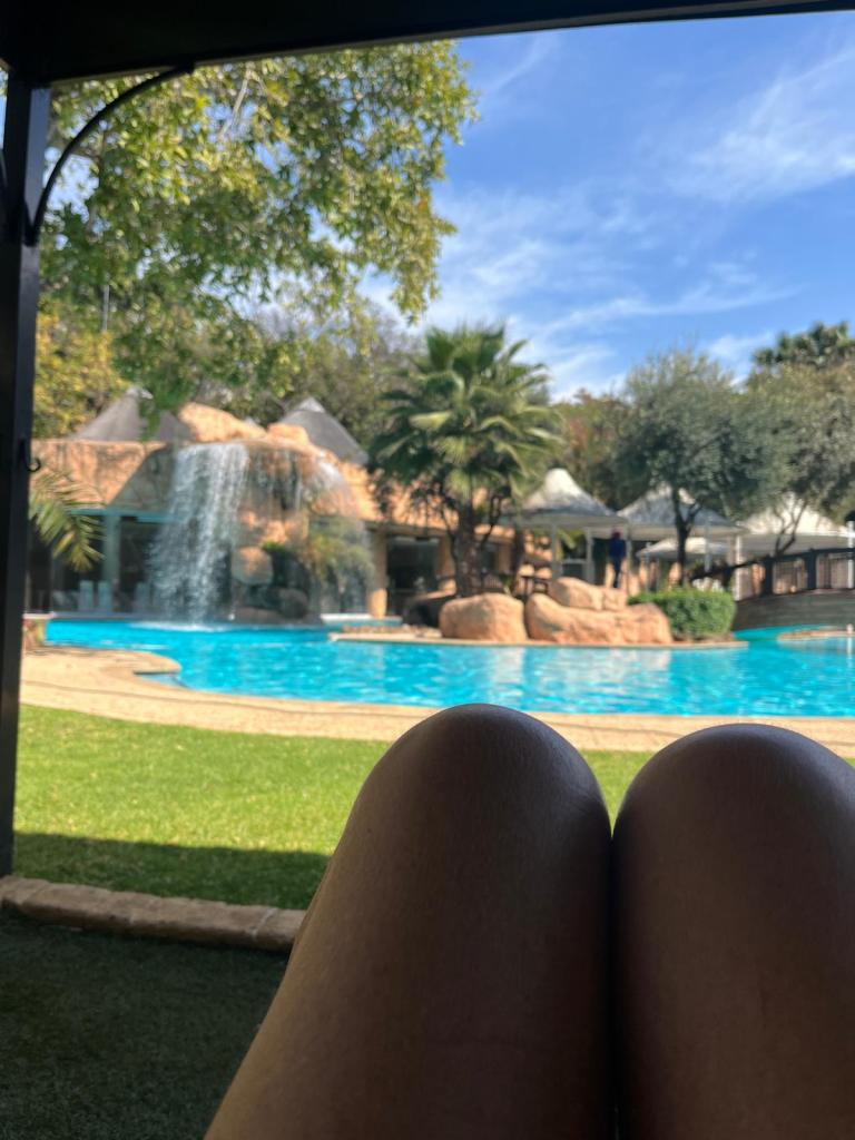 View of a swimming pool with a waterfall and palm trees in the background, legs of a person resting in the foreground.