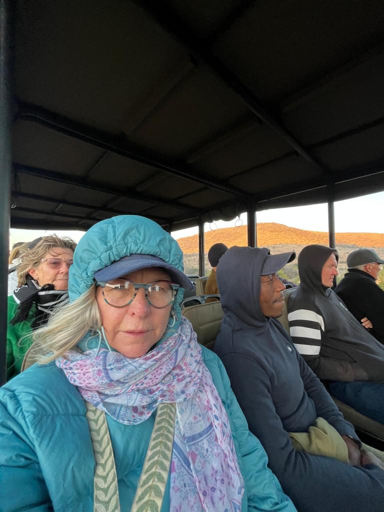 A group of people sitting in an open-top vehicle, dressed in warm clothing. The foreground features a woman with glasses and a blue jacket, alongside a man wearing a hooded top. Others can be seen in the background, some with scarves and hats.