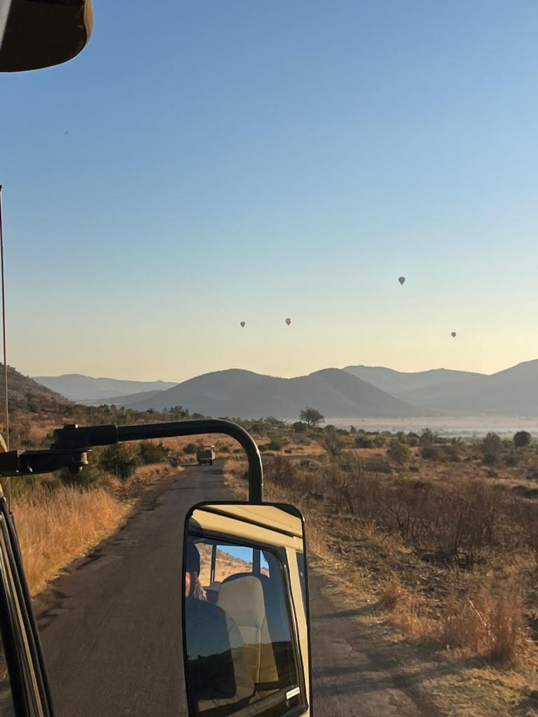View of hot air balloons in the sky over a mountainous landscape during sunrise, seen from inside a vehicle on a rural road.