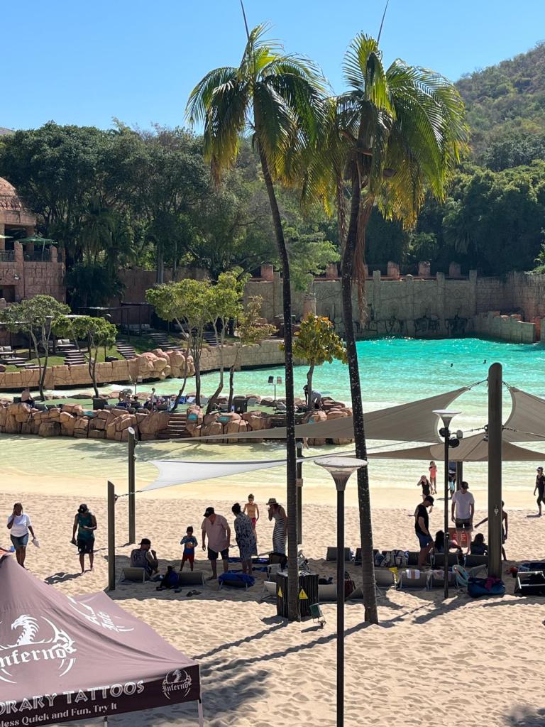 A vibrant beach scene featuring palm trees, sandy shores, and a turquoise lagoon. People are enjoying leisure activities, with some under shade structures and others playing near the water.