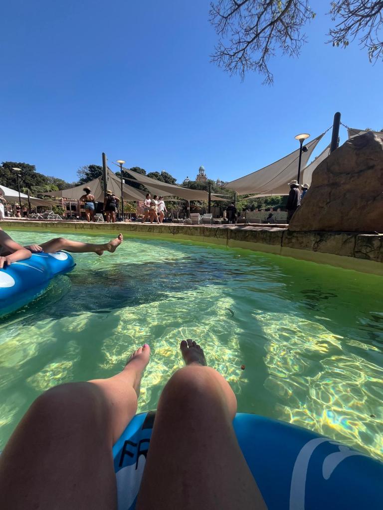 View of legs floating in a pool with clear water, surrounded by people enjoying a sunny day.