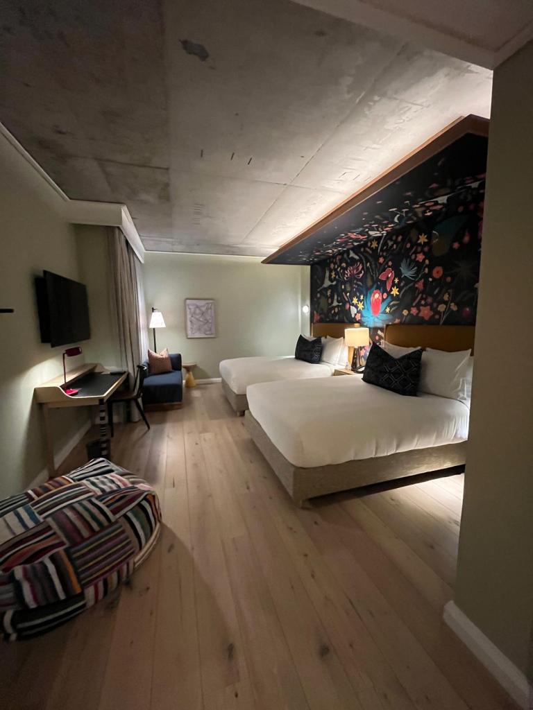 A modern hotel room featuring two beds with white linens, a colorful wall mural, a desk with a TV, and a lounge chair in a minimalistic design.
