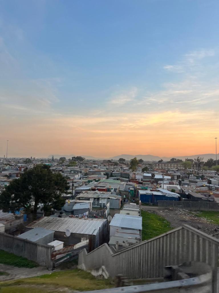 A view of a densely populated informal settlement or shantytown, featuring numerous makeshift structures under a colorful sky during sunset.
