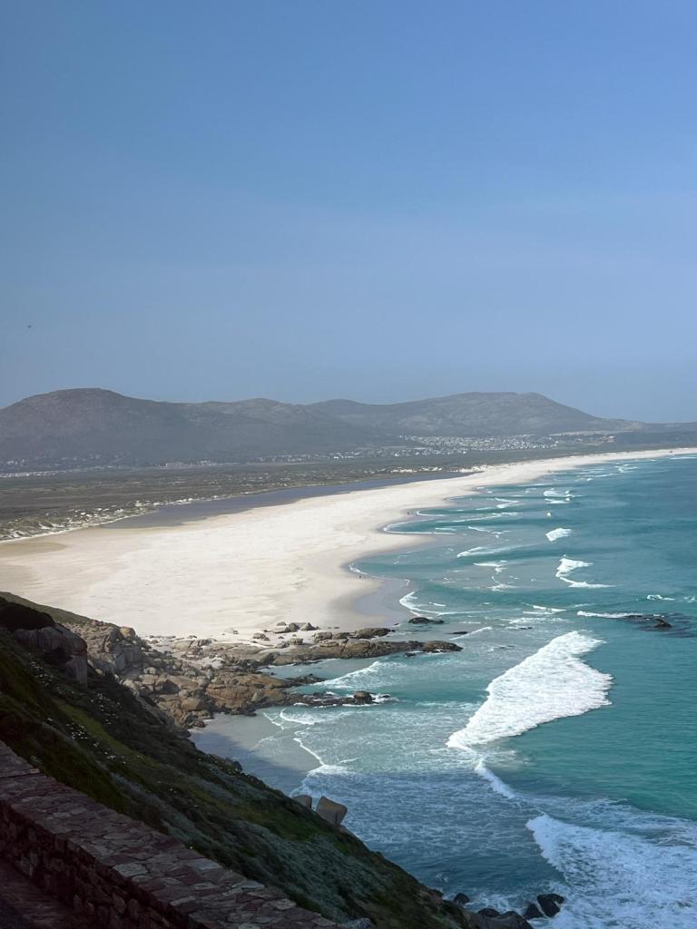 A scenic view of a sandy beach curving along a shoreline with gentle waves, framed by distant hills under a clear blue sky.