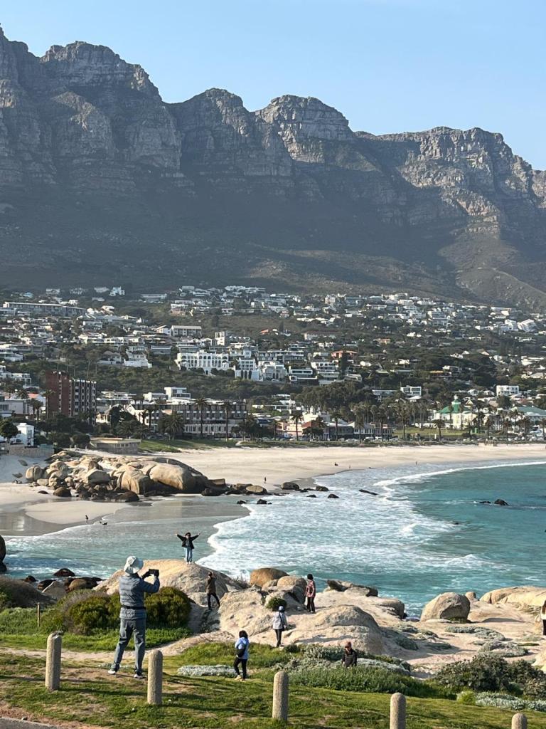 A scenic view of a beach with waves gently rolling in, surrounded by rocky formations and a backdrop of mountains. People can be seen enjoying the beach, some taking photos, while houses and palm trees line the shoreline.