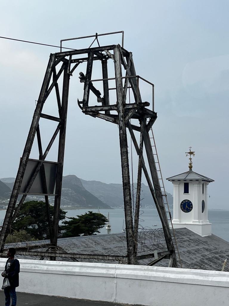 An old wooden structure resembling a bell tower with a nearby clock tower overlooking a bay and mountains in the background.