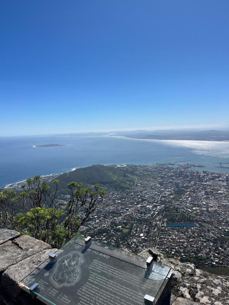 Panoramic view from a mountain overlooking a coastal city, with an island visible in the distance. A sign provides information about the area in the foreground.
