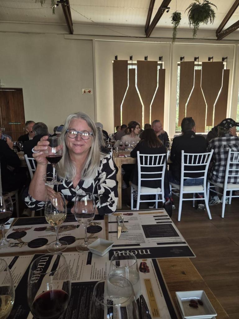 A woman with long grey hair and glasses holds up a glass of red wine, smiling at the camera. The setting is a bustling restaurant with a dining setup, featuring other patrons in the background and tables adorned with menus and wine glasses.