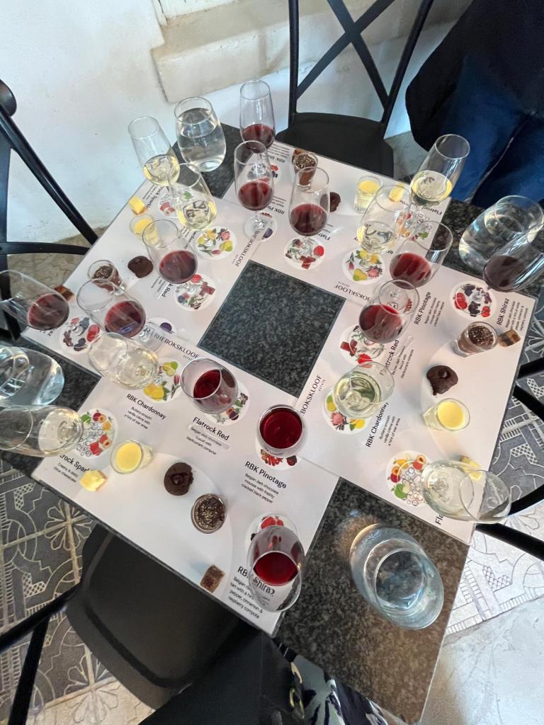 A table set up for a wine tasting event, featuring various glasses of wine, water, and tasting notes placed on the table. The table has a black granite surface surrounded by chairs.