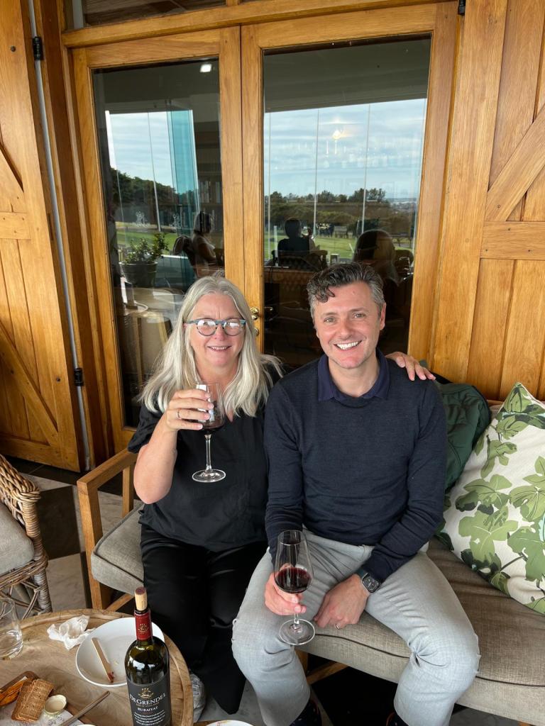 A man and a woman sitting together on a couch, both holding glasses of red wine, with a bottle of wine on the table and a cozy interior setting.