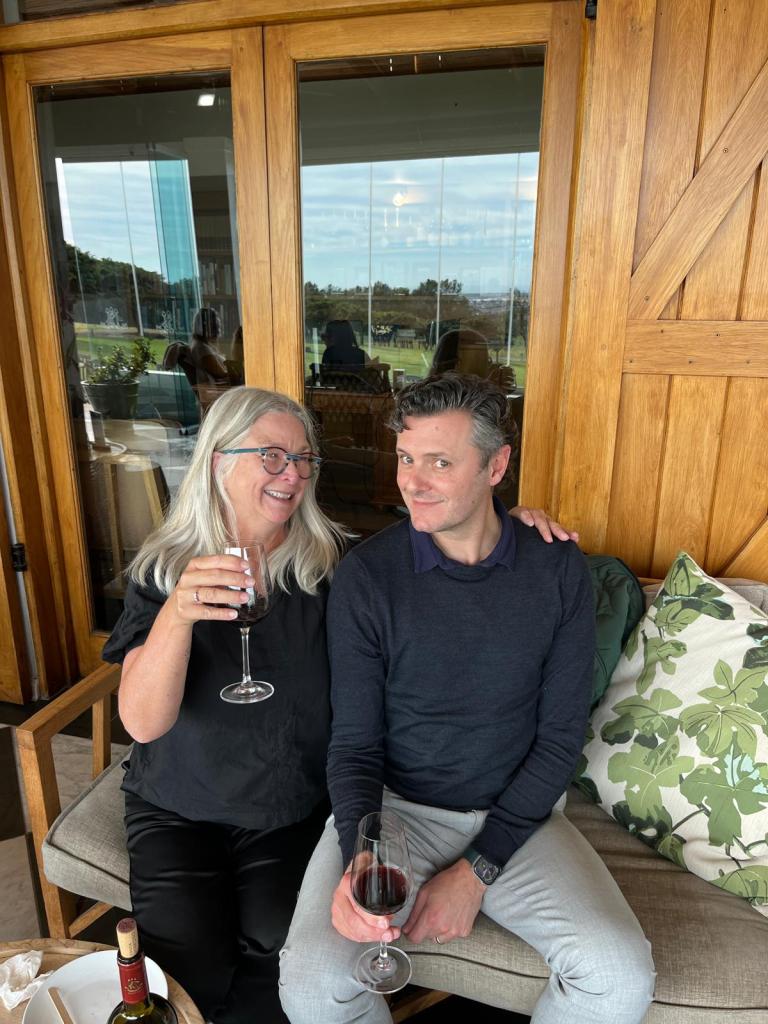 A man and a woman sitting on a sofa, both holding glasses of wine, smiling at each other. The background features large windows overlooking a green landscape.