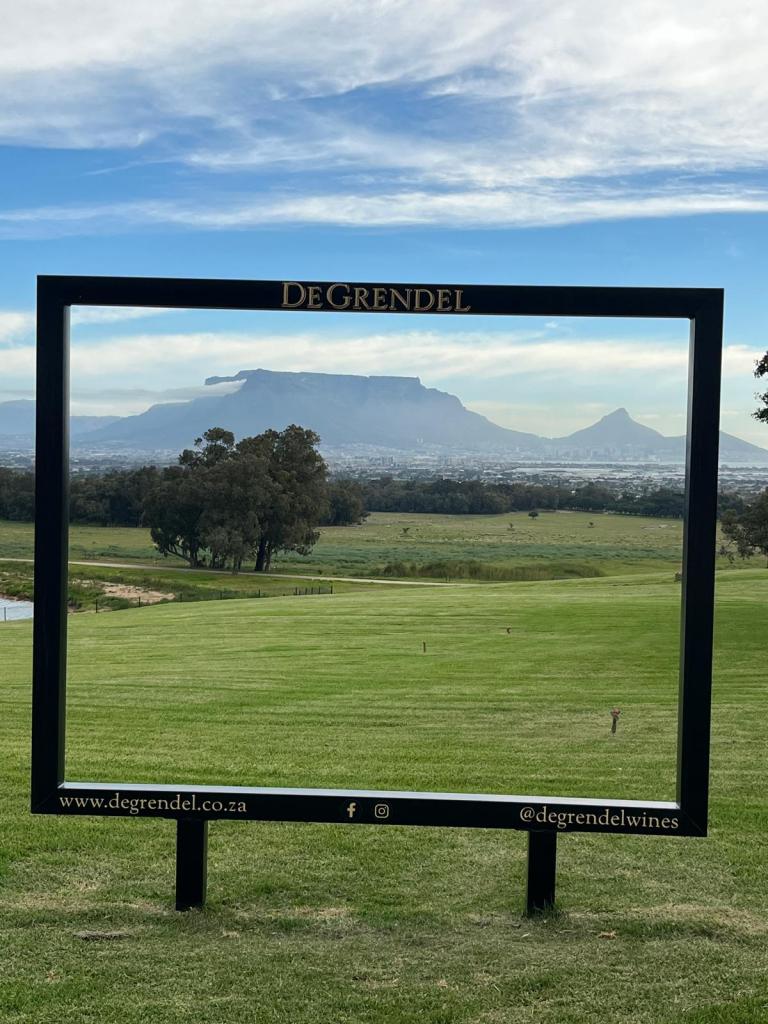 A scenic view of Table Mountain framed by a large black sign with the text 'De Grendel' at the top, set in a green landscape.