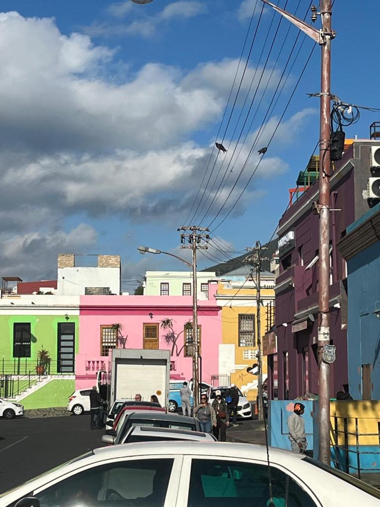 A vibrant street scene showcasing colorful buildings in shades of pink, green, and yellow, with clouds in the blue sky above. Several cars are parked along the street, and people are walking nearby.