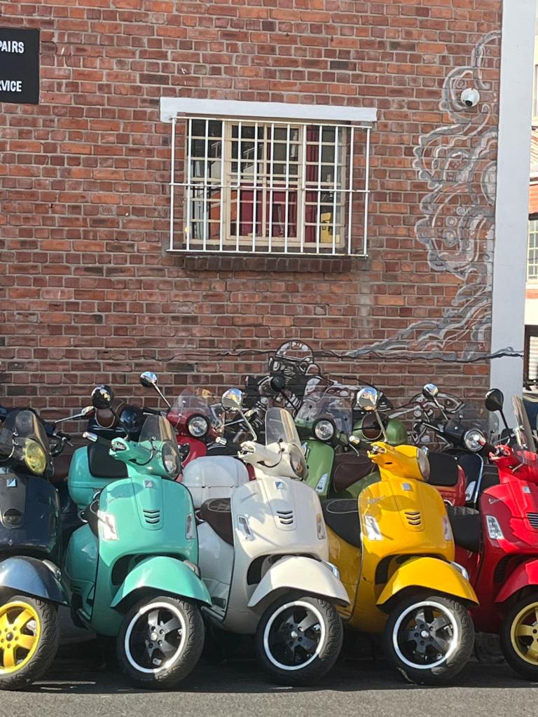 A row of colorful scooters parked in front of a brick wall featuring a barred window and part of a mural.
