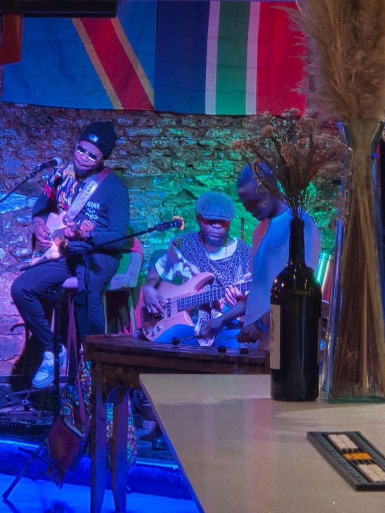 A live music performance featuring three musicians on stage, with a guitarist in the foreground, a bassist in the center, and a standing vocalist. Colorful flags hang in the background, and ambient lighting adds a vibrant atmosphere. A table with a bottle of wine and decorative elements is visible in the foreground.