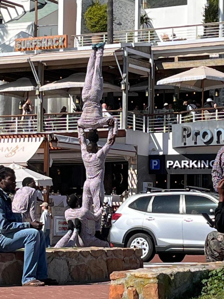 A group of street performers executing a human pyramid formation in an outdoor shopping area, with a mix of onlookers and cars in the background.