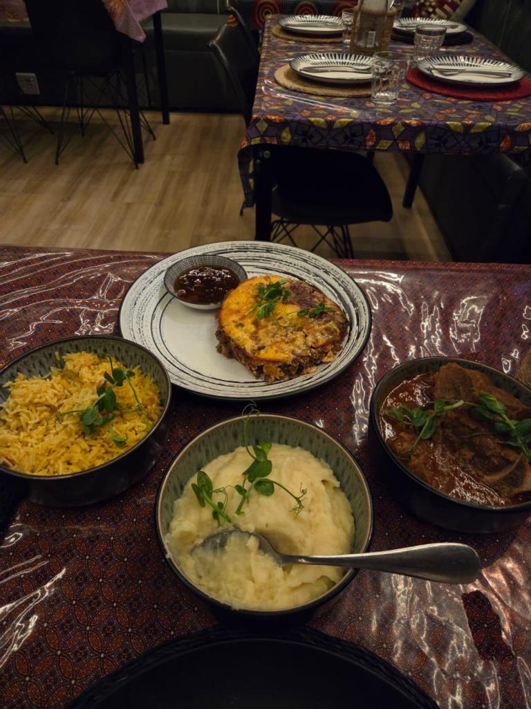 A beautifully arranged dinner plate featuring a well-cooked omelette, mashed potatoes with a spoon, flavorful yellow rice, and a bowl of rich meat stew, garnished with fresh herbs, set on a colorful tablecloth.