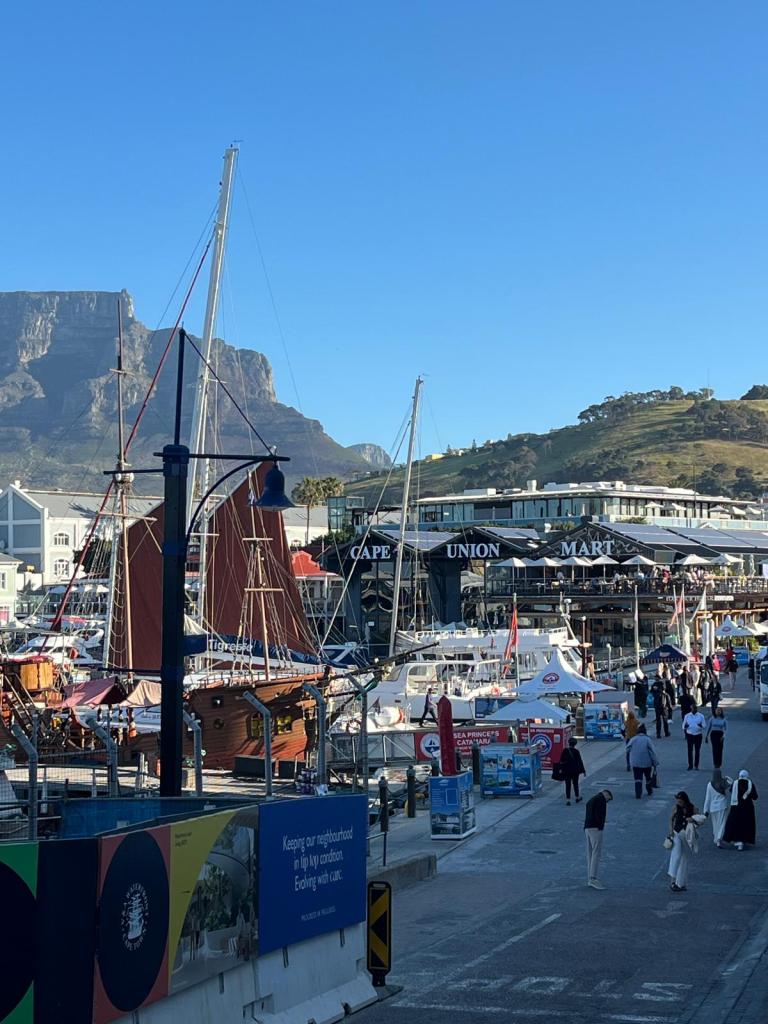 View of the waterfront in Cape Town with Table Mountain in the background, featuring various boats and people walking along the promenade near the Cape Union Mart.