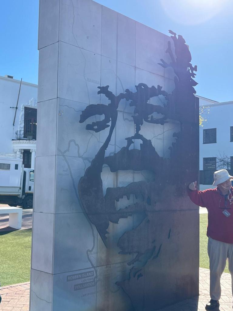 A steel monument featuring a stylized portrait engraved into its surface, with a map of Robben Island visible in the background. A tour guide stands next to the monument, explaining its significance.