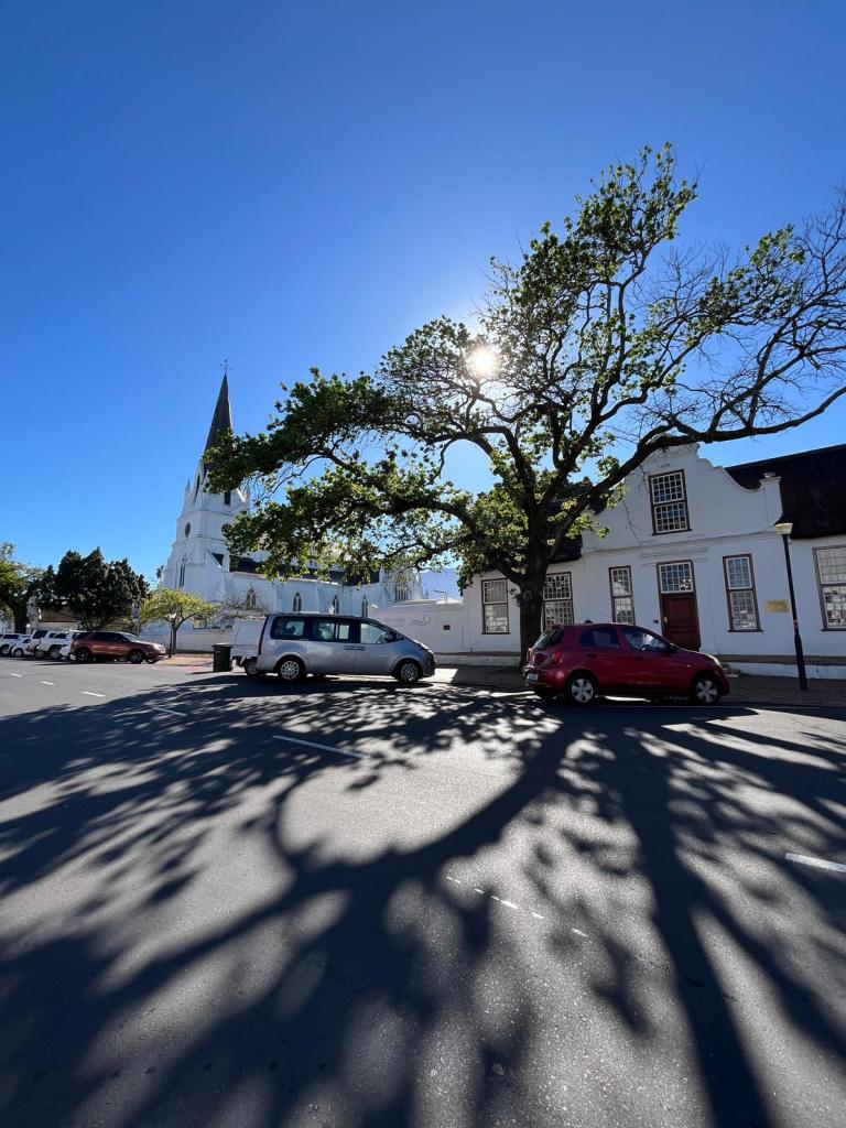 A sunny day scene featuring a church with a tall steeple, a large tree casting shadows on the street, and parked vehicles on a quiet road.