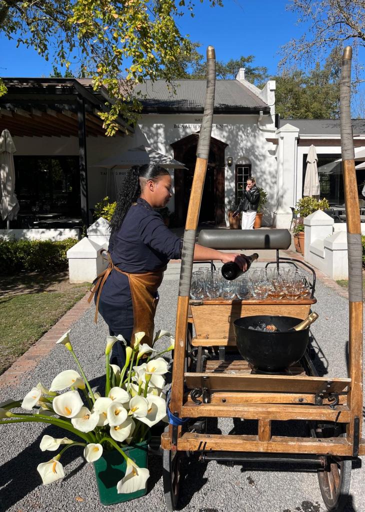 A staff member pouring a drink from a bottle into a glass on a wooden cart, with a bouquet of white flowers in a pot nearby. The scene is set outdoors in front of a restaurant under clear blue skies.