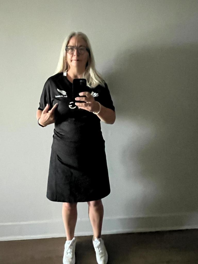 A person with long gray hair wearing glasses, a black shirt, and a black skirt stands in front of a mirror, holding a phone to take a selfie.