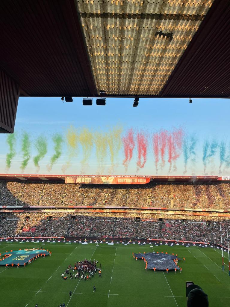 A vibrant rugby stadium filled with spectators, featuring colorful fireworks in the sky and teams gathering on the field.