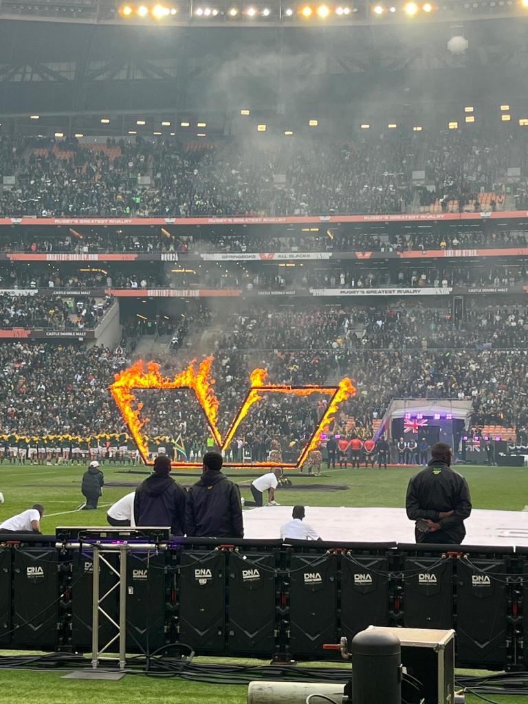 A sports stadium filled with a cheering crowd, featuring a fiery display creating the shape of a 'V' on the field, with performers and staff in the foreground.