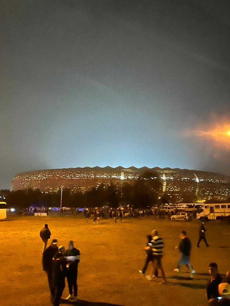 A stadium illuminated at night with a crowd of people gathered in the foreground, set against a hazy sky.