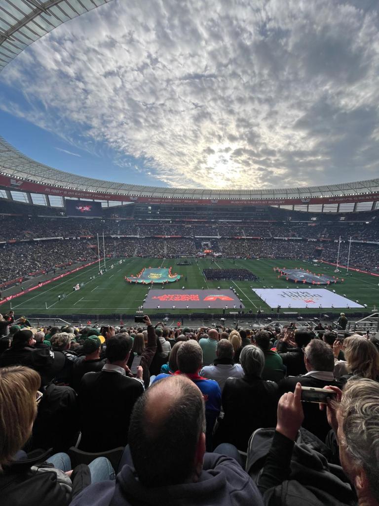 A panoramic view of a rugby stadium filled with spectators, featuring a grassy field with large banners and flags, under a cloudy sky.