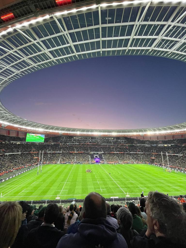 View of a rugby stadium filled with spectators during a match, featuring a large green field and dramatic sunset sky.