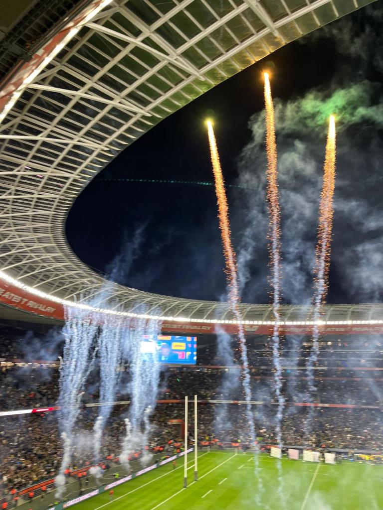 Fireworks and smoke erupt from the roof of a stadium during a rugby match, with fans visible in the stands and a large scoreboard in the background.