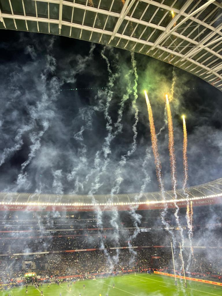 Fireworks illuminate the night sky above a packed rugby stadium, with smoke trails and a modern structure visible.