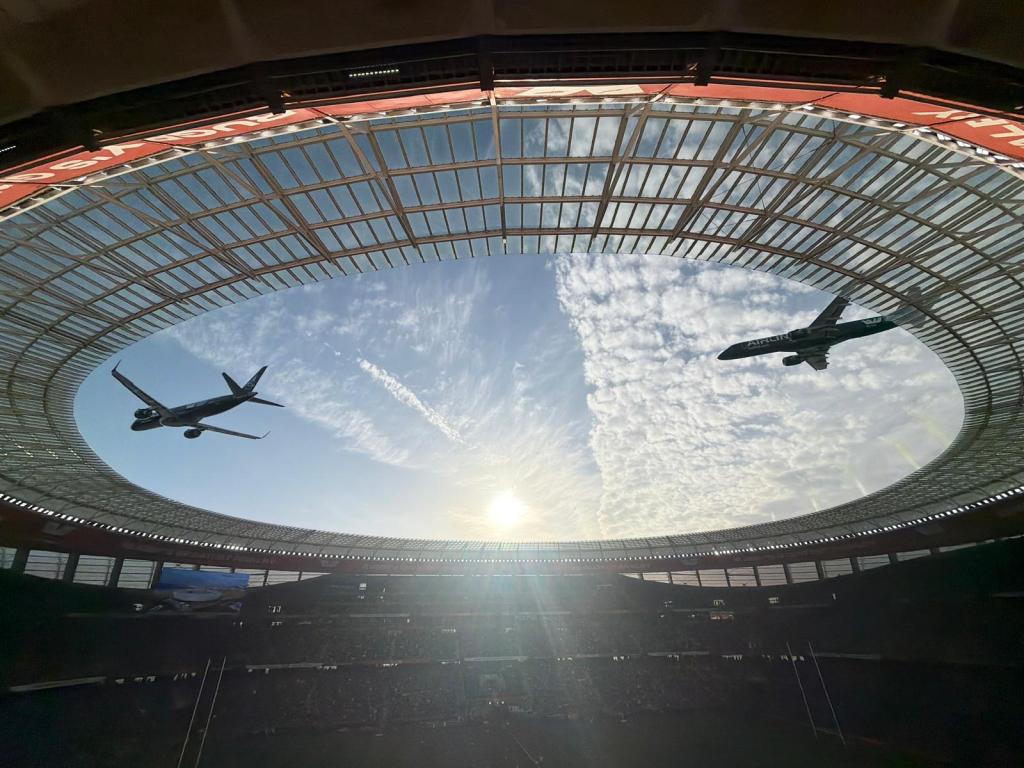 A unique perspective of a stadium with two airplanes flying overhead against a backdrop of a bright sky and clouds.