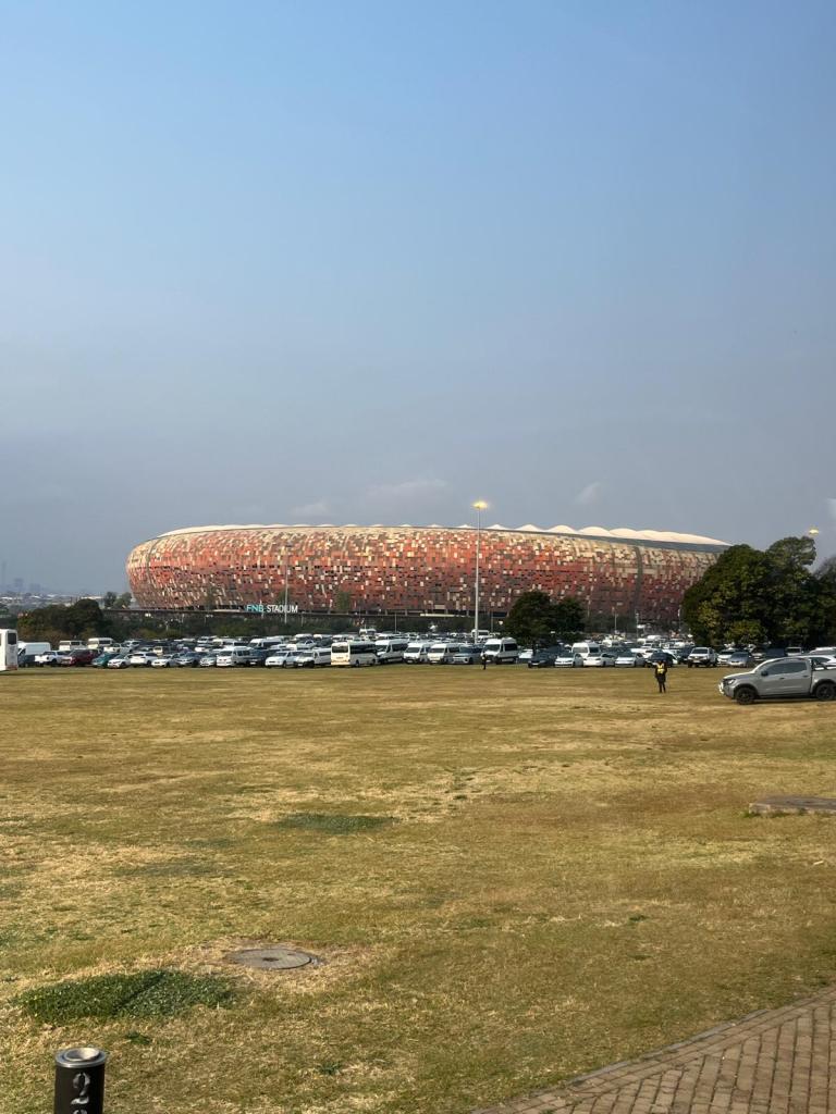 A large, modern stadium with a distinctive patterned exterior, surrounded by a grassy area and parked vehicles in the foreground.