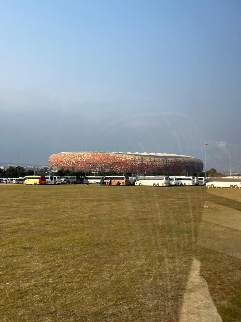 A large stadium with a distinctive design, surrounded by a grassy area and several parked buses on the side.