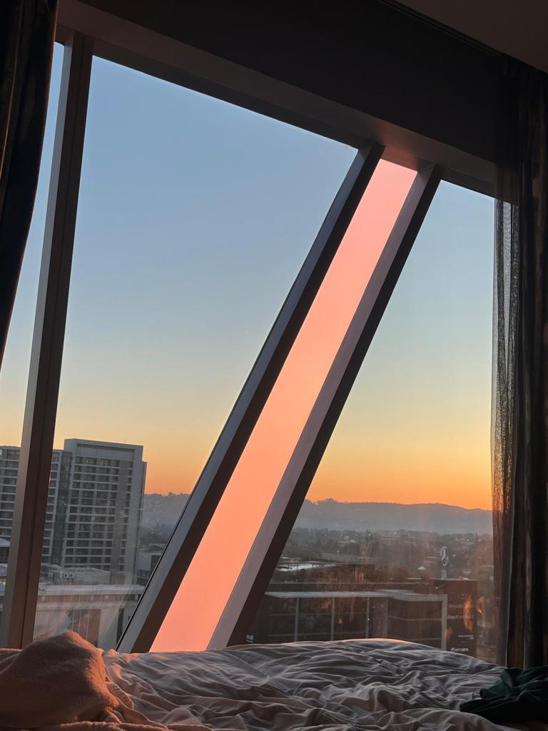 A view from an angled window showcasing a sunset with vibrant colors, framed by modern architecture, and a glimpse of a cityscape below.