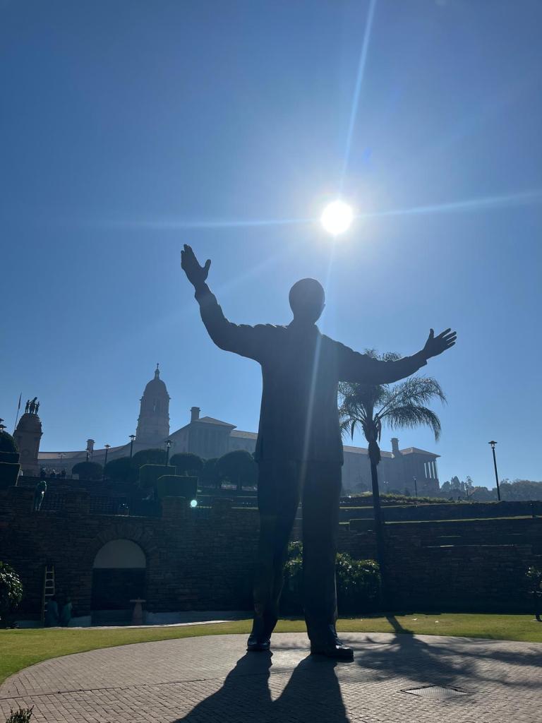 Silhouette of a statue with outstretched arms against a bright sunlight, surrounded by greenery and historical buildings in the background.