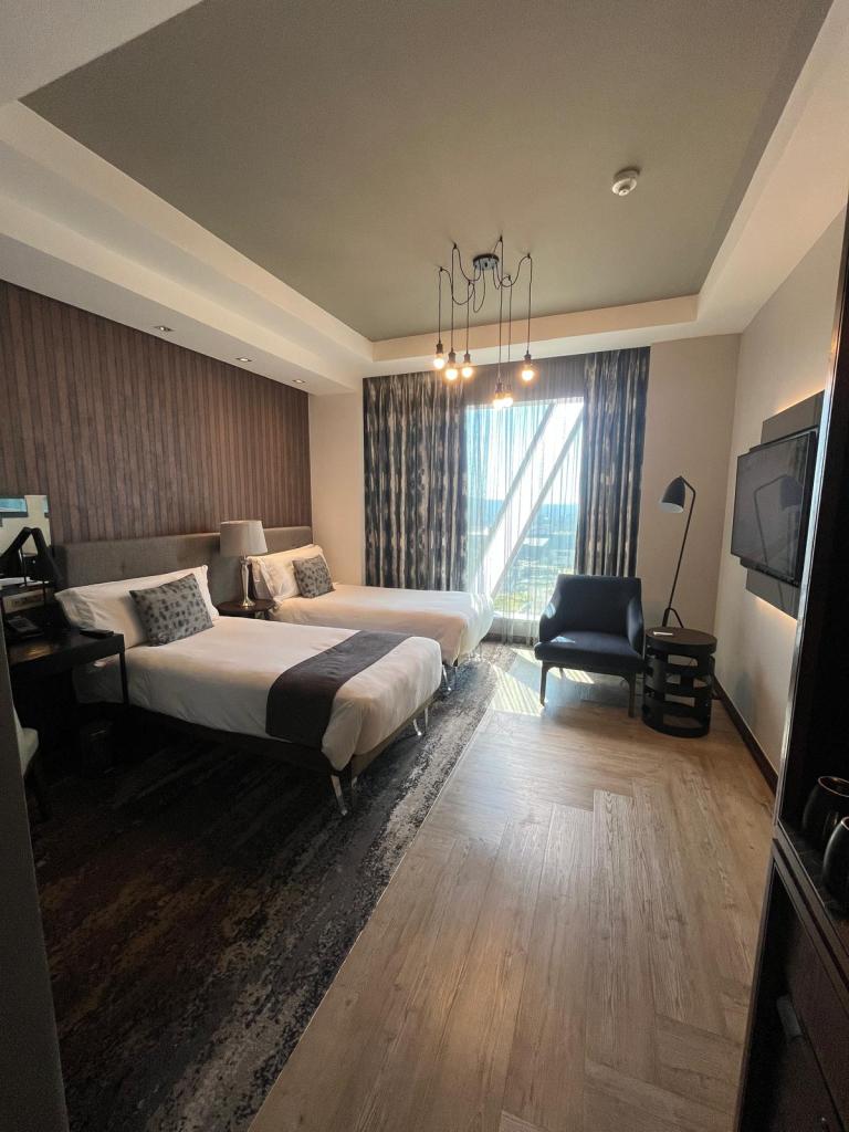 Modern hotel room with two beds, a chair, desk, and large window with curtains, featuring stylish decor and wooden flooring.