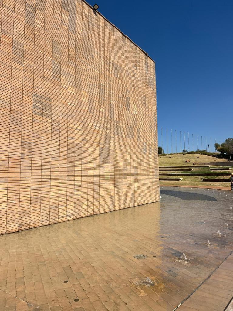 A large brick wall with a smooth texture, partially reflecting sunlight, adjacent to a tiled surface with water features, under a clear blue sky.