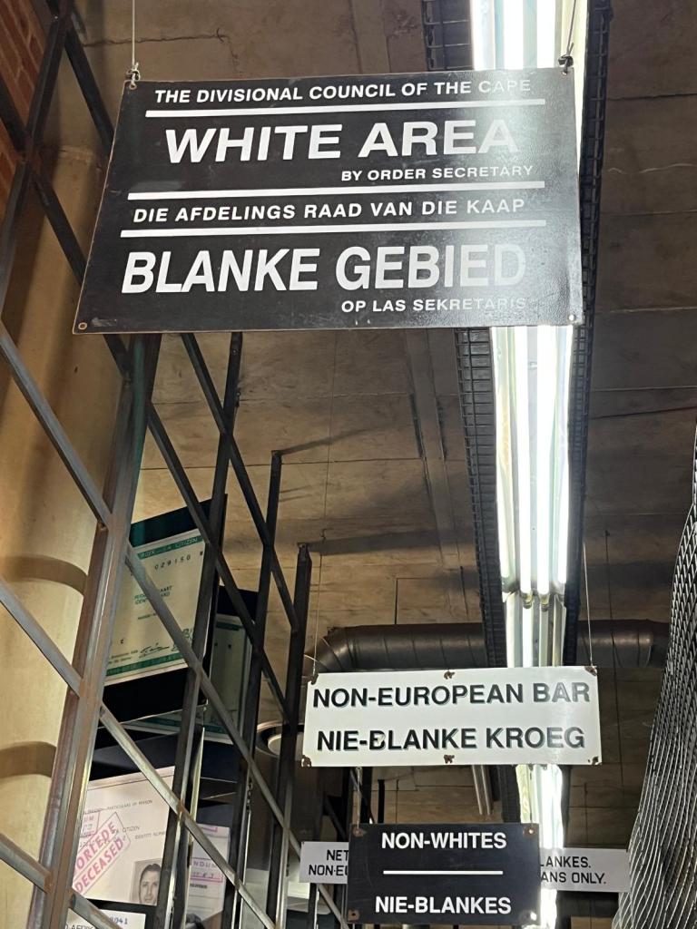 Interior view showing signs labeled 'White Area', 'Non-European Bar', and 'Non-Whites' in both English and Afrikaans, indicating segregation policies.
