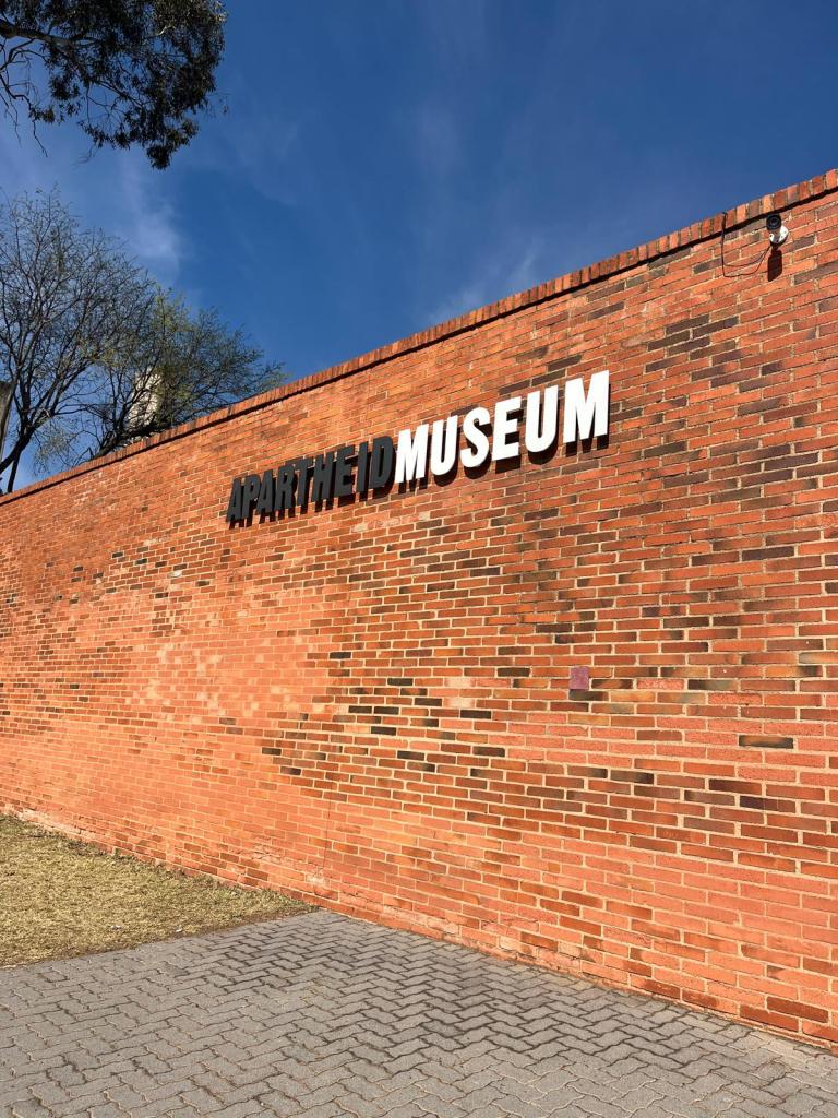 Exterior view of the Apartheid Museum's brick wall with the museum's name prominently displayed in bold white letters against the red brick.